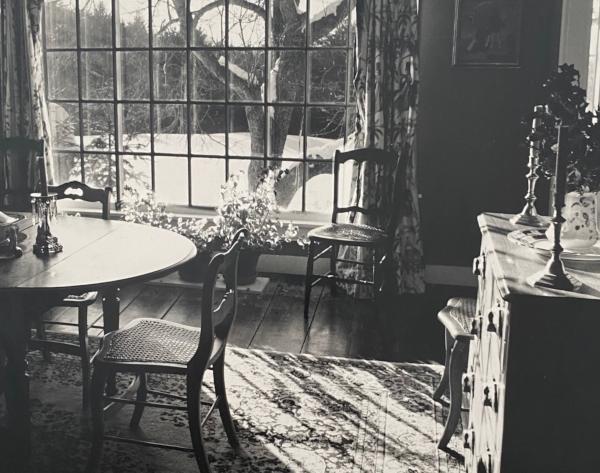 Vermont Light is a black and white photograph of a dining room filled with antiques with a large window overlooking a snow-filled field and an apple tree. The light from the window saturates the room