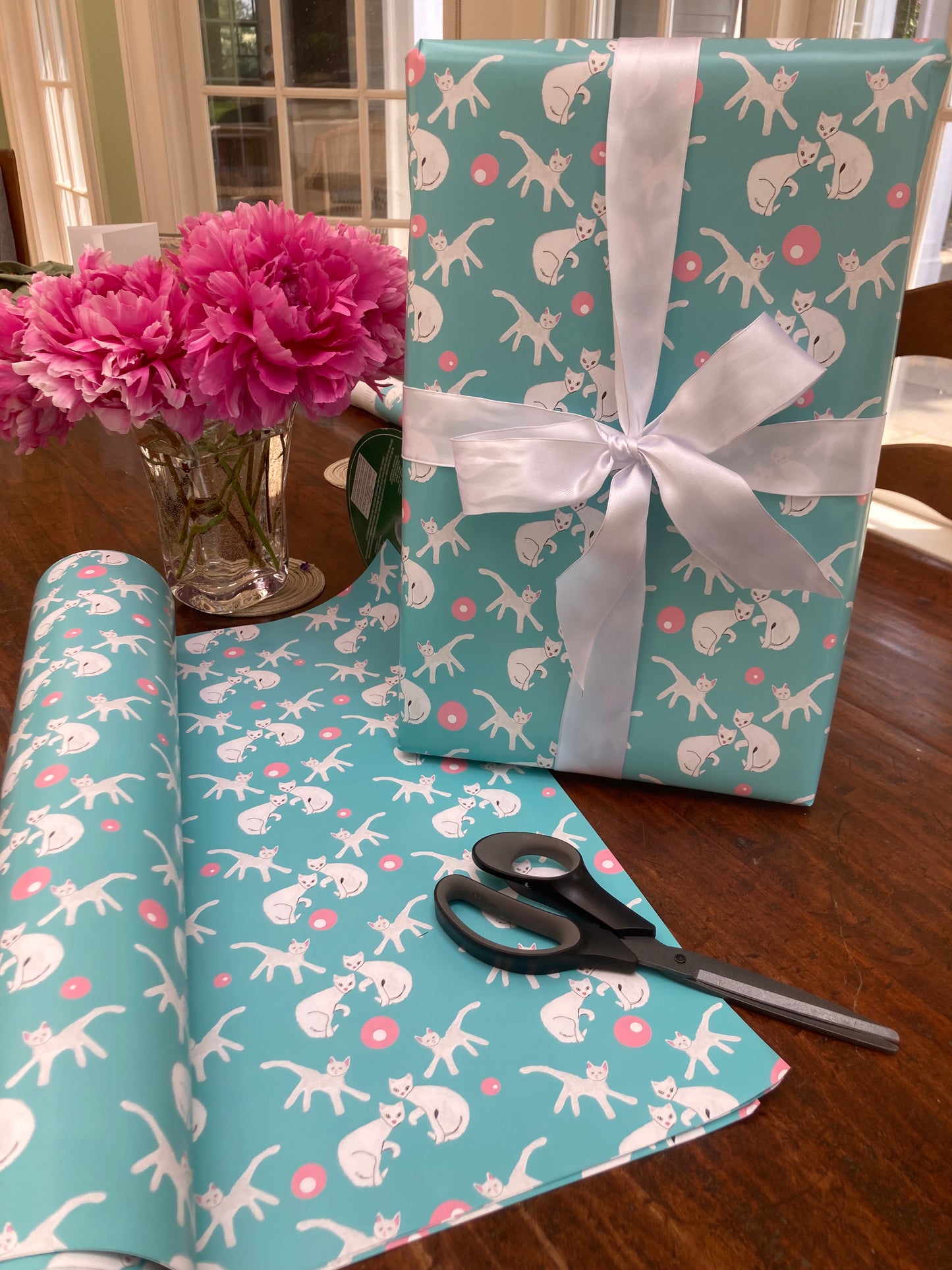 Mockup of the Fancy Felines wrapping paper with sheets of gift wrap unfurled on table top with a shirt box wrapped with a ribbon and standing on end to display the gift wrap. A vase of pink peonies and a pair of scissors are also on the table.