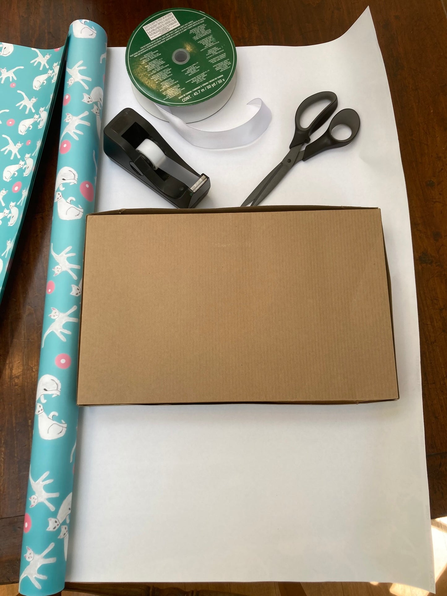 photo of roll of wrapping paper titled Fancy Felines Wrapping Paper unfurled under a brown shirt box about to be gift wrapped. There is a roll of ribbon, a tape dispenser and a pair of scissors to give idea of size of the roll of the gift wrapping.