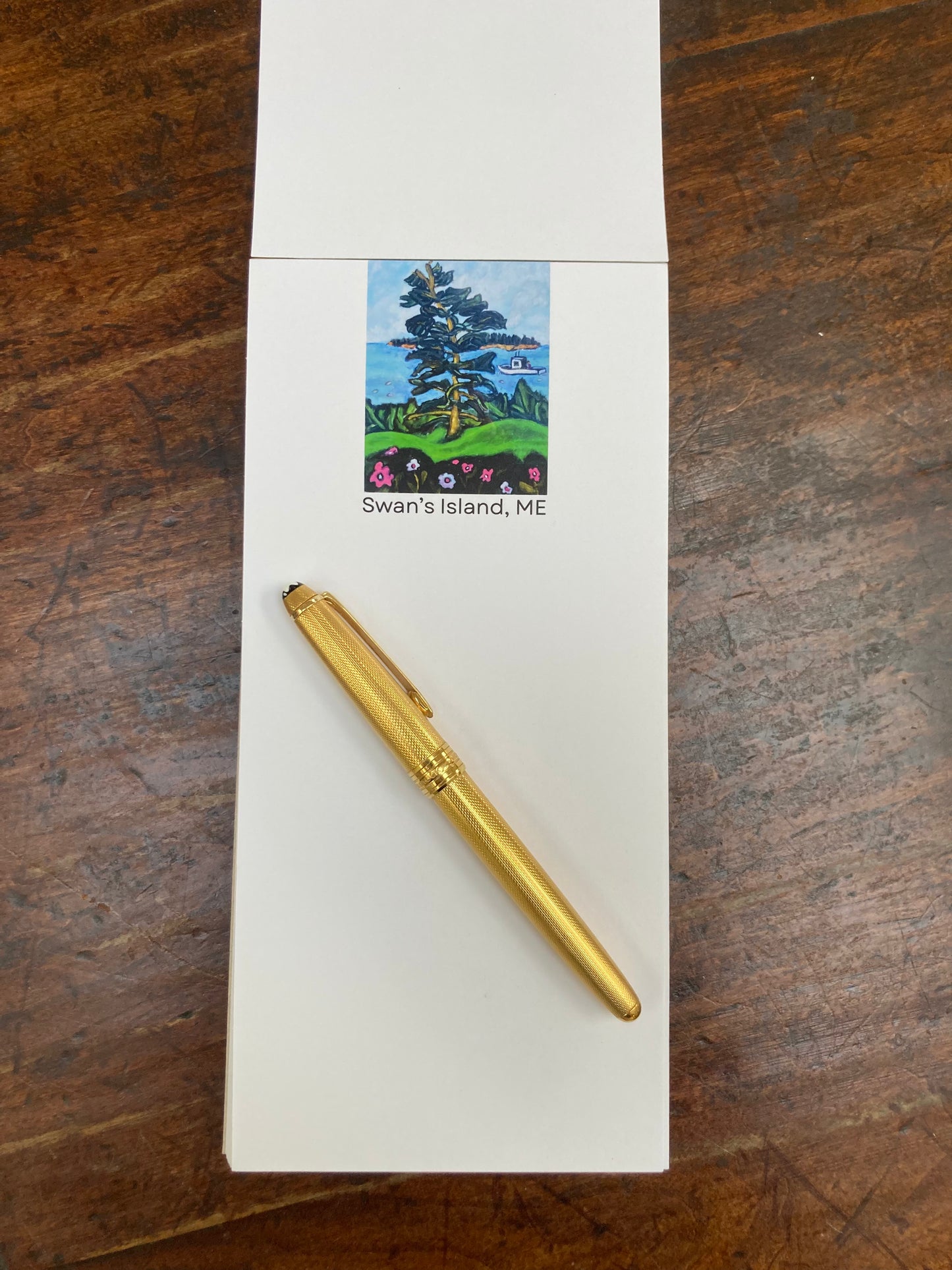 Swan's Island Notepad flat on brown tabletop with gold pen across pad beneath the picture.