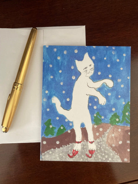 photograph of the winter themed greeting card titled Caspar On Skates. The white cat on blue background card is lying on a brown desktop with white envelope and gold pen.