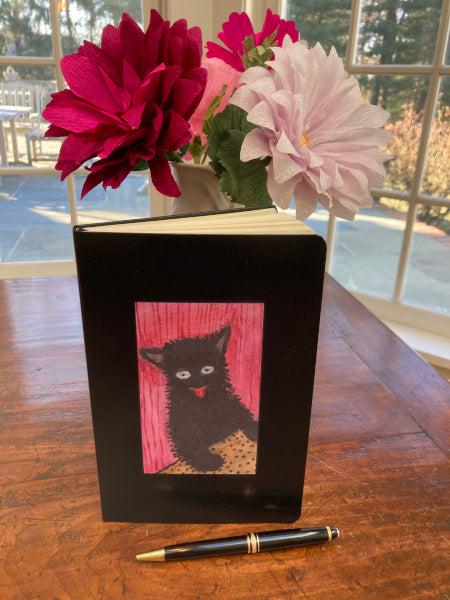 Mock up of Happy Cat journal in black is propped upright on a wooden dining table with a vase of colorful flowers behind that accentuate the deep red background of the image.