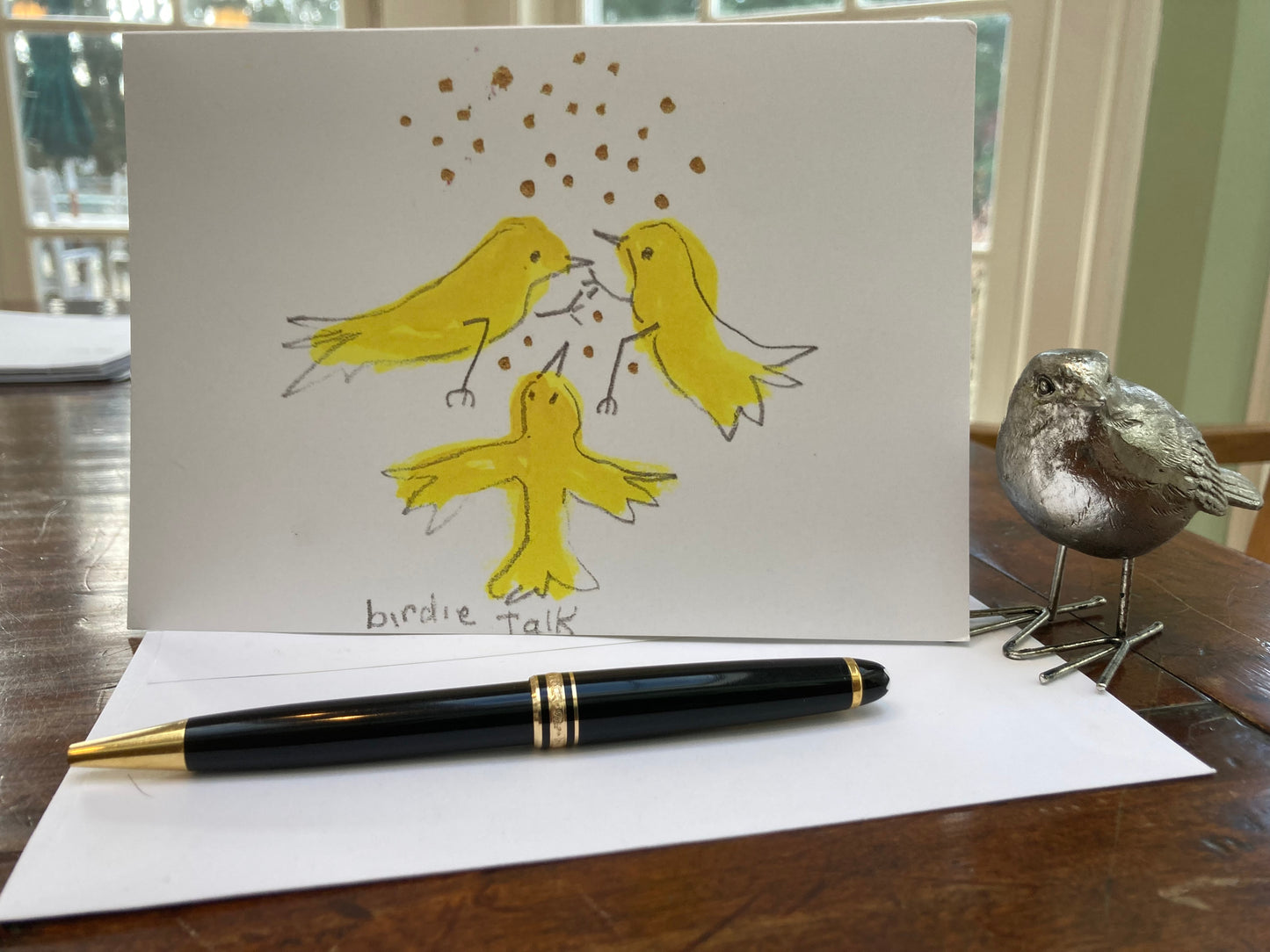 The front of the greeting card Birdie Talk displayed on brown desk top with envelope, black pen, and metal figurine of a song bird to the right.