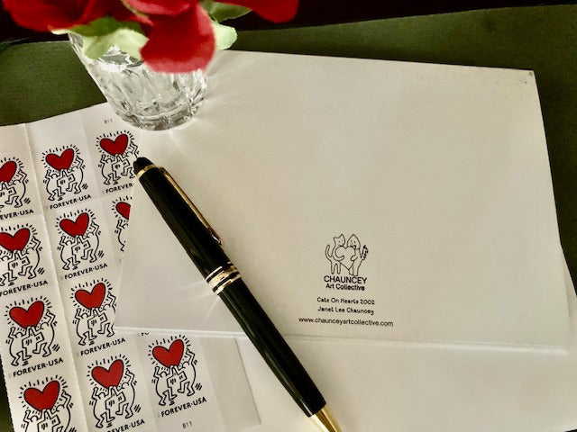 Back of the greeting card Cats On Hearts displayed on a green desk blotter with white envelope, black pen, partial view of sheet of valentines day postage stamps and cut glass vase with red roses.