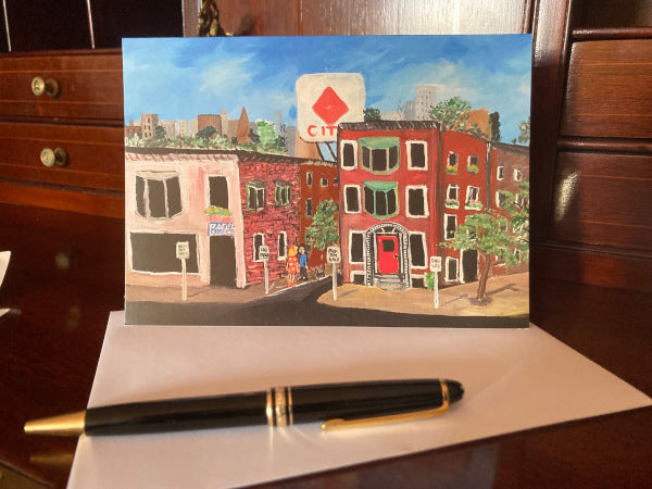 Mockup of the card as it is propped up on a wooden desk with the street scene facing the camera. The envelope and a fountain pen are also shown.