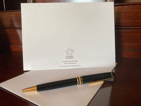 The back of the card Caspar Brings Gift displayed standing upright on a brown desk with a white envelope and a black pen in front.
