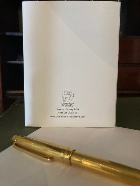 Photograph of the back of the autumn greeting card entitled Vermont Turkey which is standing upright on a desk top with logo and title in view along with a white envelop and a gold pen.