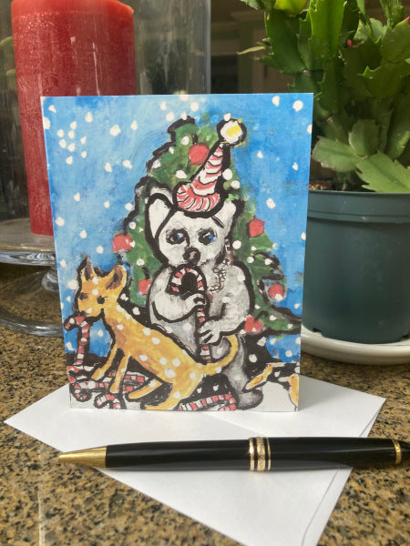 Photograph of the holiday greeting card titled Astonishing Creatures With tasty Treats that shows the card standing upright on a granite surface with envelope and black pen. Red candle in hurricane and green Christmas cactus partially visible in the background.