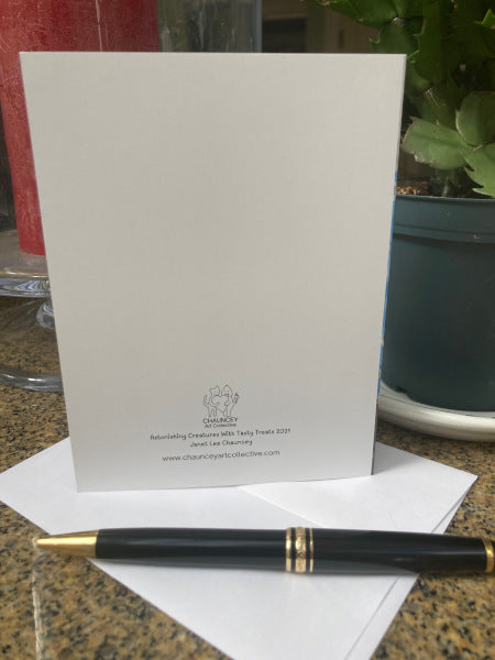 Photograph of the back of the holiday greeting card entitled Astonishing Creatures With tasty Treats. The card is shown standing upright on a granite surface so you can read the logo and title, a white envelope is seen along with a black pen.