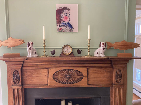 Jeanette acrylic print portrait is seen here hanging centered on the wall above a brown pine mantle with various objects on the mantle including a clock, two Staffordshire dogs, two rust colored metal birds and two wooden fish.