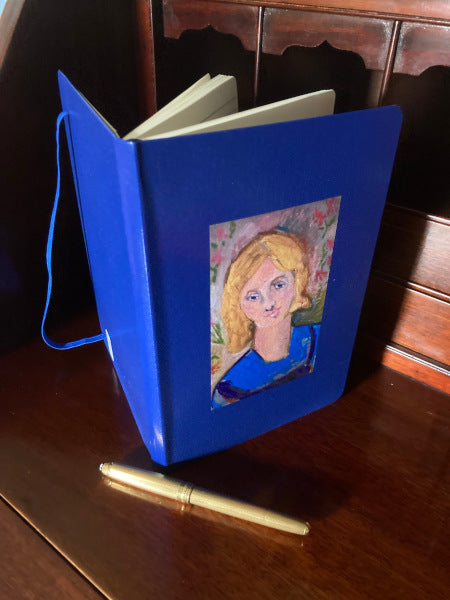 Mockup of Yellow Haired Girl journal in blue atop a wooden desk with the pages fanned open.