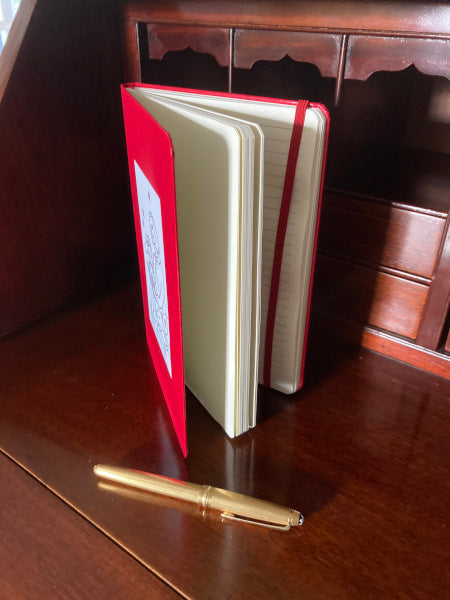 On Being Idolized red journal book mockup standing on a wooden desk with its pages fanned out.