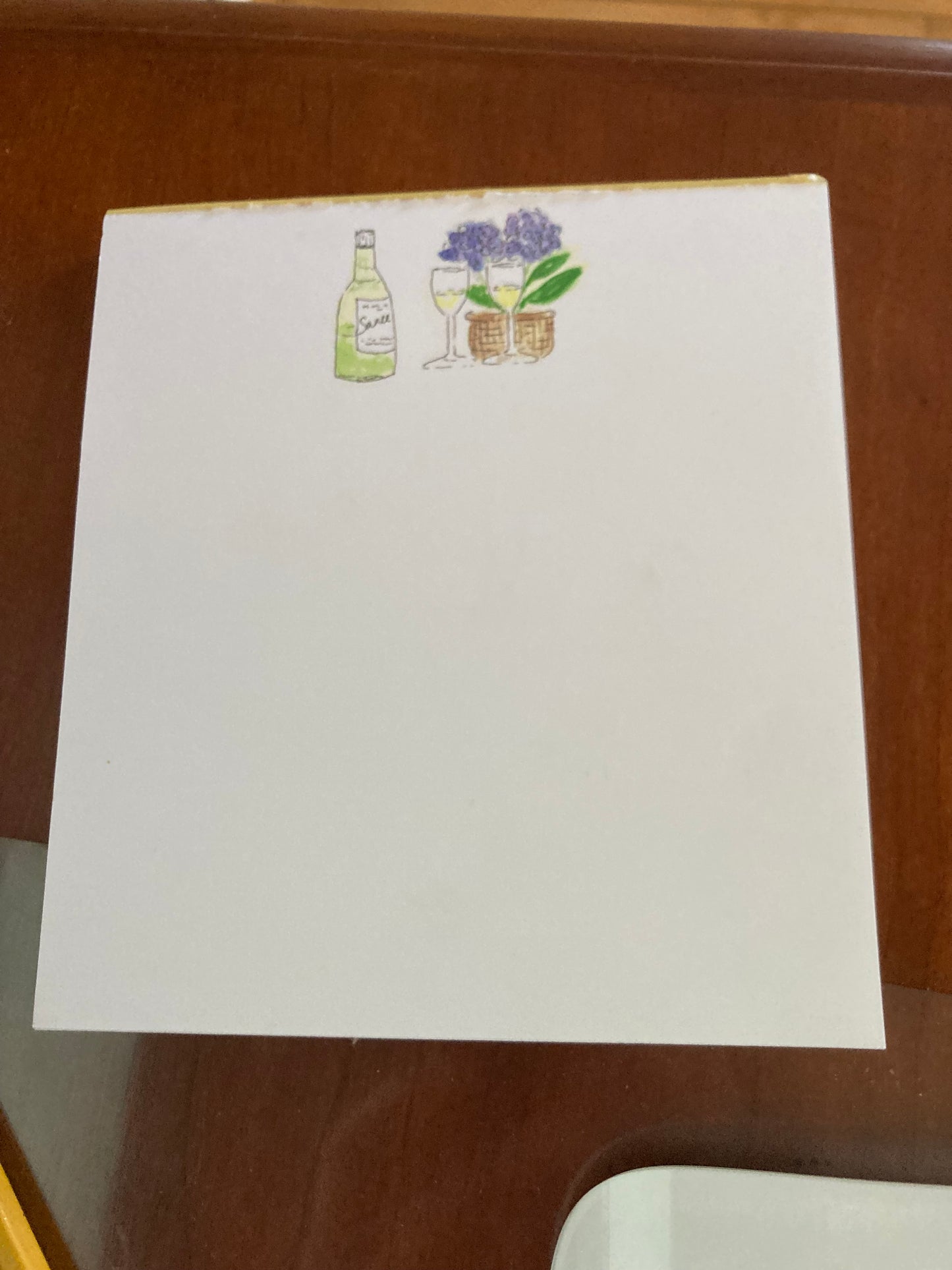 Photograph of Wine And Hydrangeas notepad on brown desktop. The notepad has illustration of green wine bottle with two filled wine glasses to the right and in front of a wicker basket with blue hydrangeas and green leaves. The image is centered on the white paged pad.