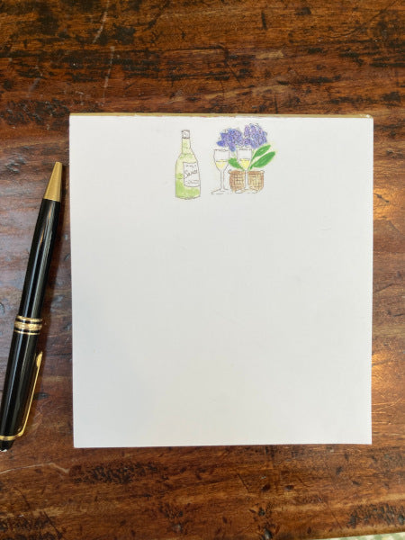 A wine bottle stands beside a basket of blue hydrangeas with two wine glasses on this Wine And Hydrangeas notepad. The pages of the notepad are white and the images are centered on the top third of the pad. black pen lies on the brown desktop beside the pad.