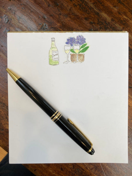 View of a notepad titled Wine And Hydrangeas atop brown desk. Image on notepad is of wine bottle, two wine glasses and basket of hydrangeas centered on top quarter of page with black pen lying on top to give perspective.