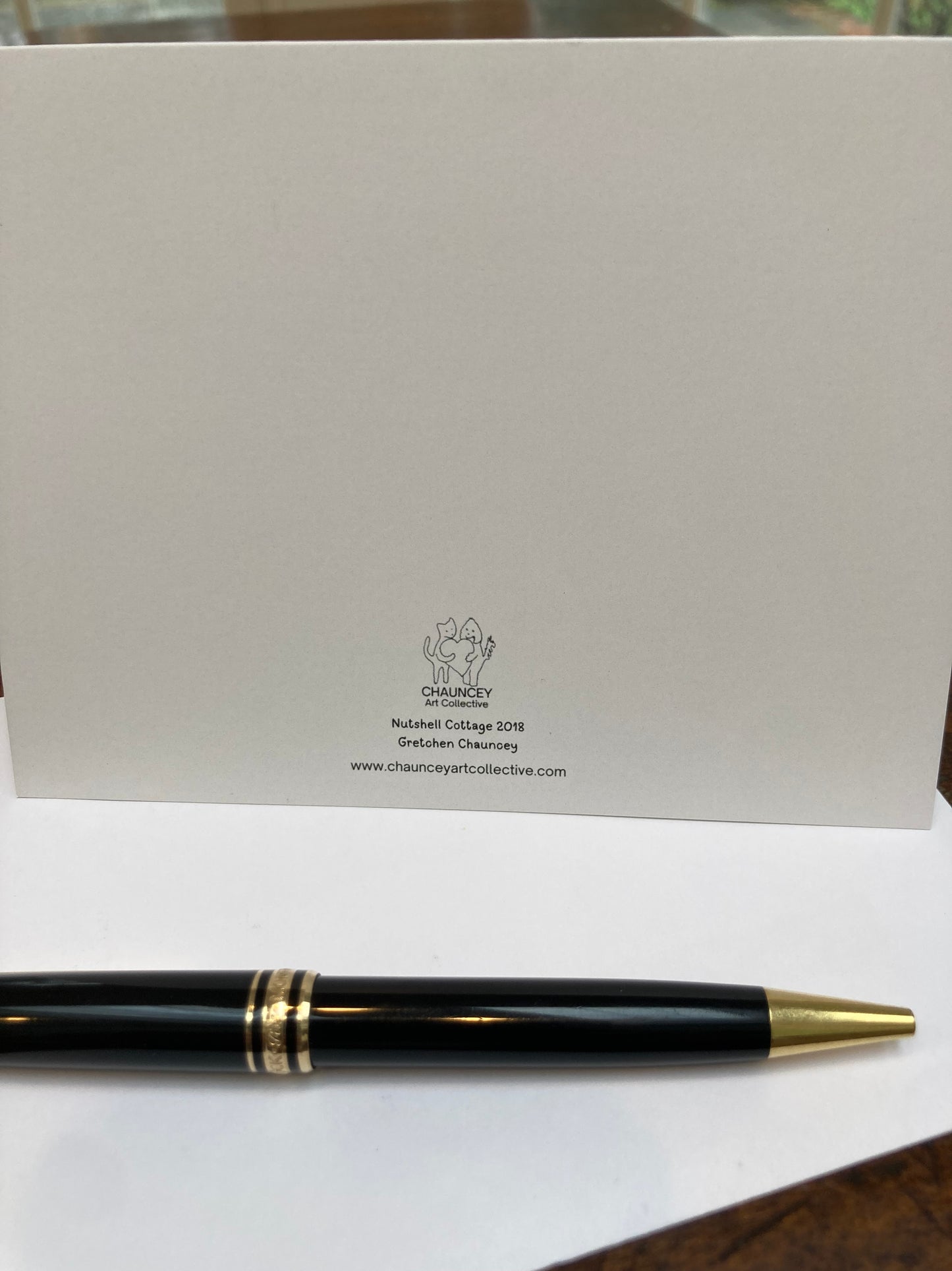Mockup of the back of the card with the title Nutshell Cottage and the Chauncey Art Collective logo. Set on a wooden table with a fountain pen.