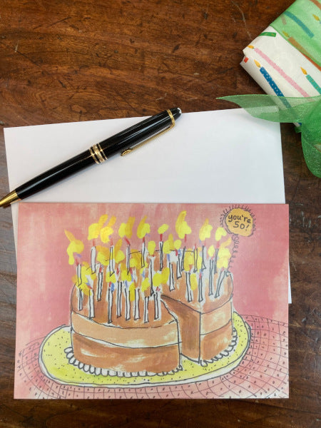 The front of the birthday card You're 50! displayed on a wooden table with the envelope, a wrapped present and a fountain pen.