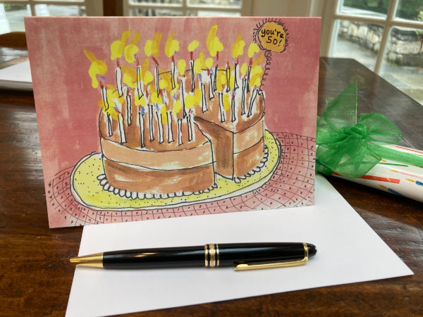 The front and envelope of the birthday card You're 50! on a wooden table with a wrapped gift with a green ribbon.