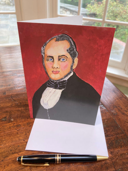Mockup of the front of the greeting card, "The Good Doctor" standing upright on wooden table with white envelope and black pen. The image is of a stately gentleman wearing black tie and black jacket with white shirt. He has blue eyes and faces forward for his portrait.
