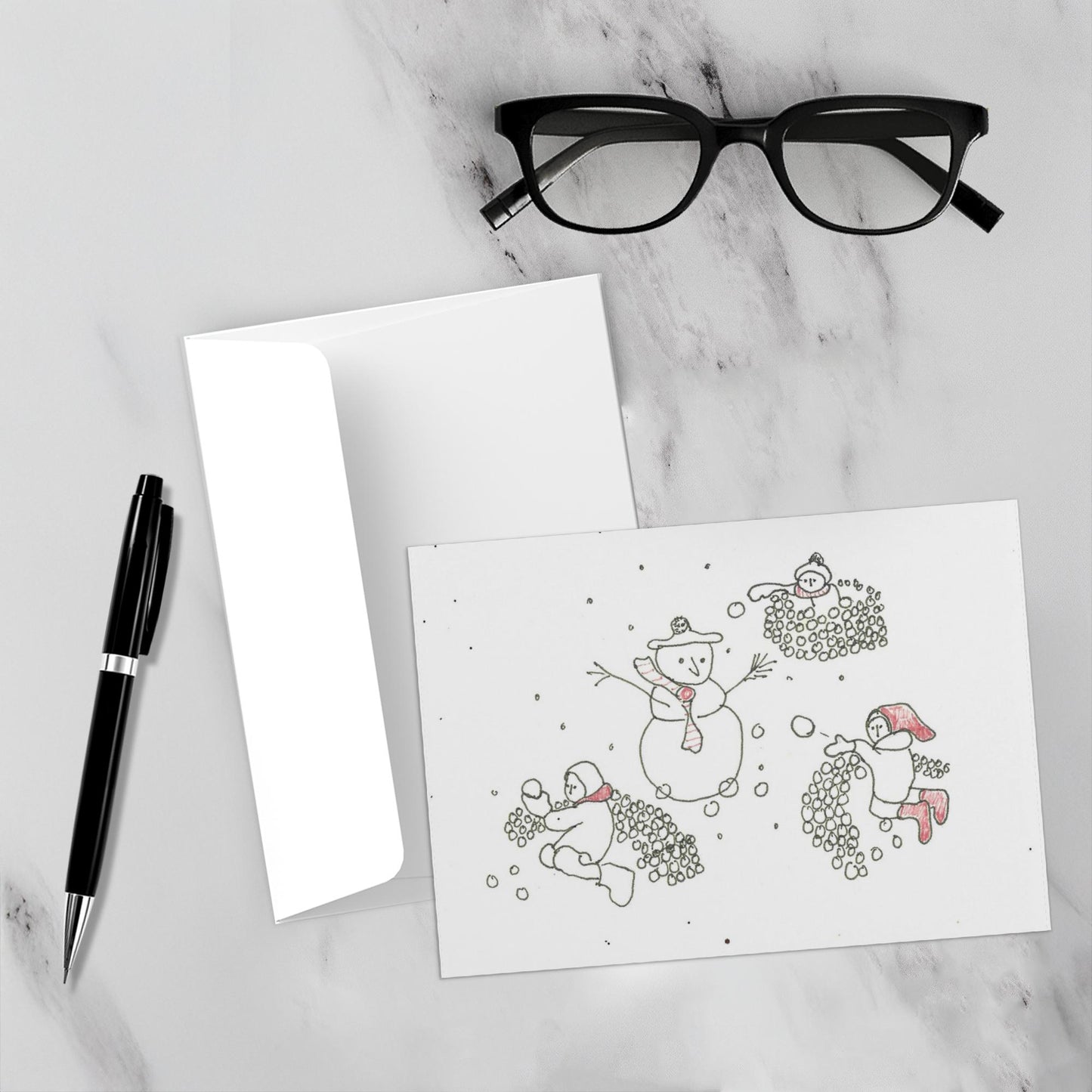 Mockup display showcasing the front of the greeting card Snowball Fight With A Snowman lying on a gray marble surface along side a white envelope, a pair of black reading glasses and a black pen.