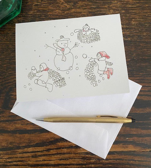Front of the Snowball Fight With A Snowman greeting card displayed lying flat on brown desktop with white envelope and gold pen.