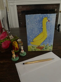 A Few Snowflakes, Yellow Duck With Flower greeting card is displayed on a brown desktop beside some easter ornament figurines, and easter basket and in front is a white envelope and a gold pen.