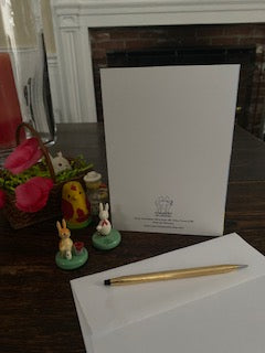 The greeting card A Few Snowflakes, Yellow Duck With Flower is displayed on a brown table top alongside a small easter basket with red flowers and some easter figurines as well as an envelope with a gold pen.