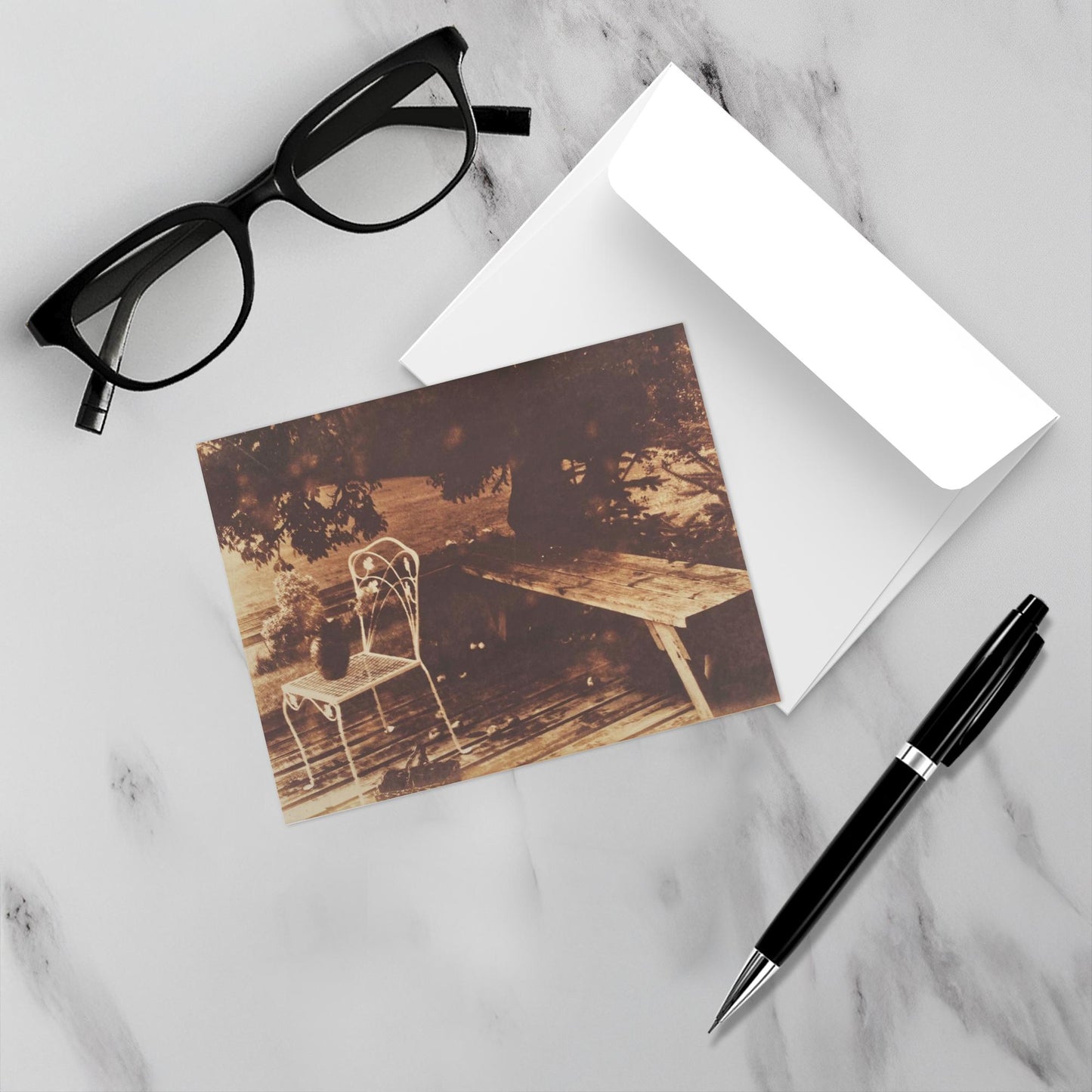 Mockup of the card on a white marble backdrop accompanied by the envelope, reading glasses and a pen.
