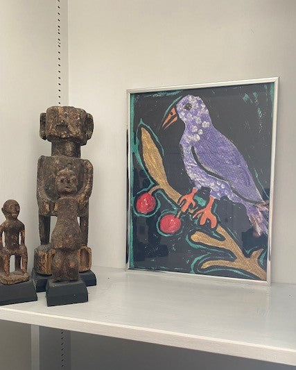 A framed Birdie on a Twig art print sits on a shelf alongside some African sculptures. The colors of the art print really pop against the white background.