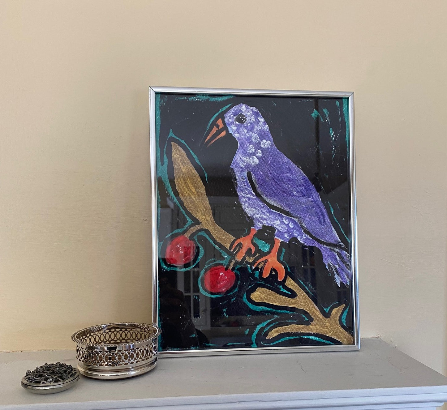 Birdie On A Twig art print is framed in a silver frame on a mantle next to some silver objects. The orange of his feet and his purple body look adorable against the black background.