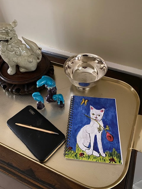 A mock up of the cobalt blue spiral notebook with the cat with a rose in it's mouth is resting on a gold tray by a white foo dog statue, a silver bowl and a golden pen.