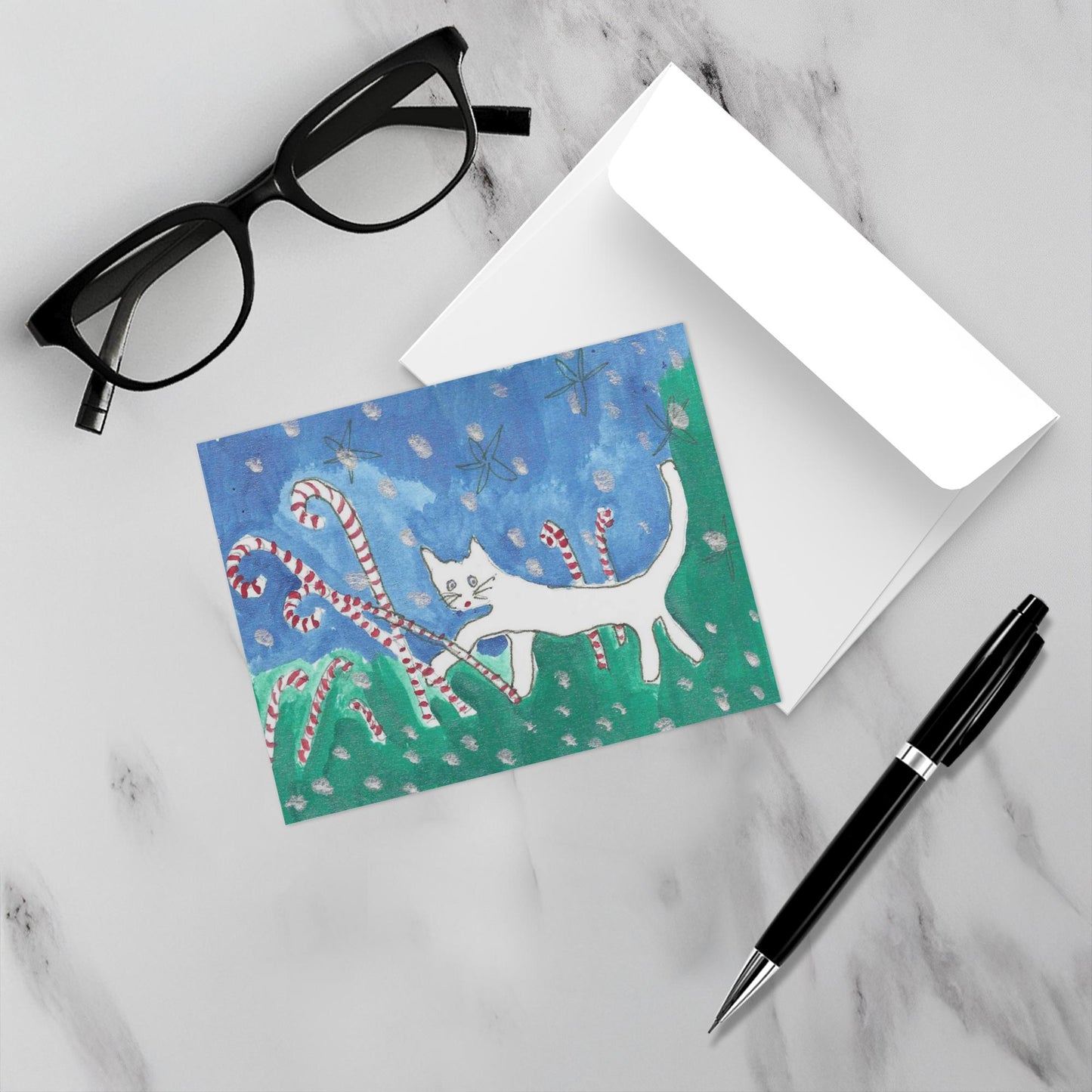 Mockup of the holiday card Caspar In Candyland which shows the front of the card with the cat among candy canes, a white envelope, a pair of reading glasses and a black pen.