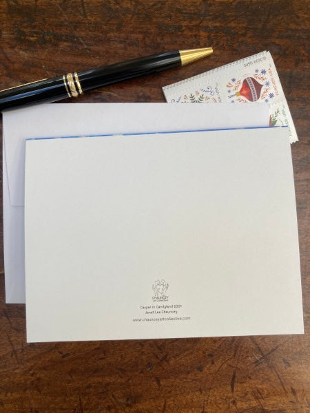 Photograph of the back of the Christmas card titled Caspar in Candyland which shows the card with Chauncey Art Collective logo lying on wooden desk with a partial view of an envelope and holiday postage stamps and a black pen.