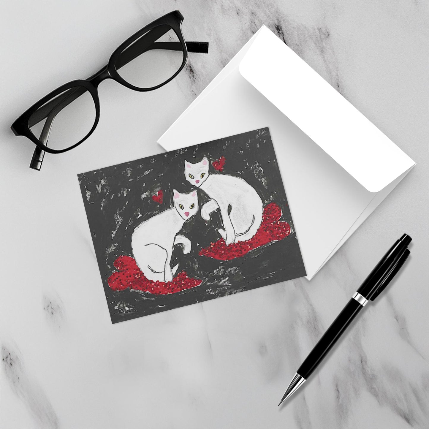 Mockup display showcasing the front of the greeting card Cats On Hearts lying on a gray marble surface along side a white envelope, a pair of black reading glasses and a black pen.