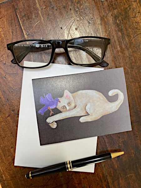 Mockup of Cat With Bird notecard laying on a wooden table with a pen and reading glasses.