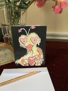 Photograph of the greeting card titled Crazy Heart Bird standing on end with white envelope and gold pen in the foreground. In the background is a cut glass vase with pink flowers and partial view of a gold desk clock.