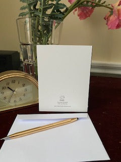 Photograph of the back of the greeting card Crazy Heart Bird see standing upright on desktop with cut glass vase of pink flowers in background and envelope with gold pen in the foreground. Partial view of a gold desk clock is to the left.