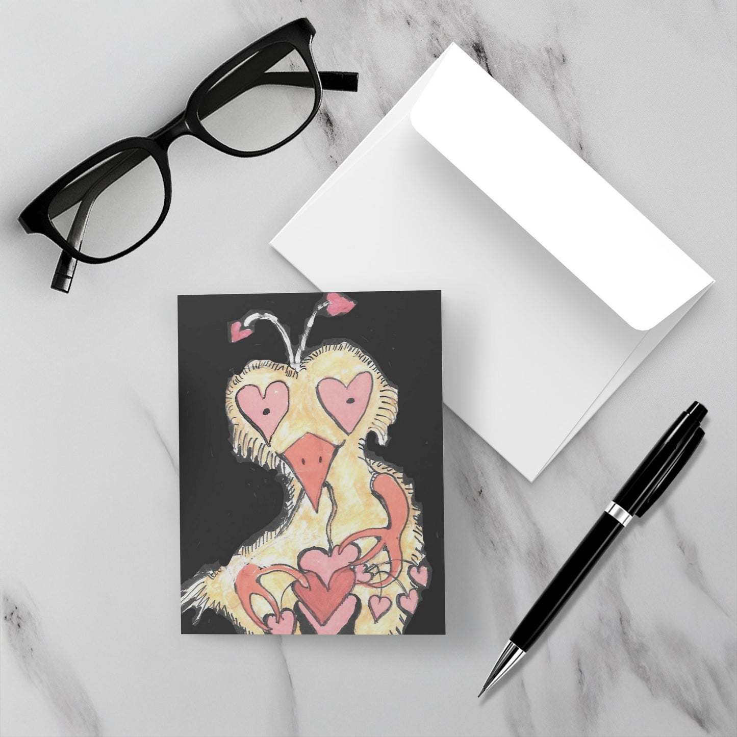 Mockup of the greeting card Crazy Heart Bird which shows the front of the greeting card with the yellow bird lying atop the white envelope on a grey marble topped surface with a pair of reading glasses on the upper left and a black pen on the lower right.
