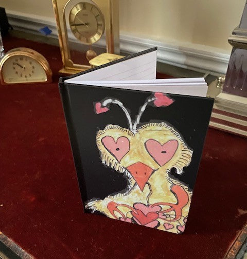 Photograph of the front of the Crazy Heart Bird journal standing upright on red velvet surface of desk. the journal is slightly open so you can see the lined pages within. Some objects are in the background partially seen among them two gold desk clocks.