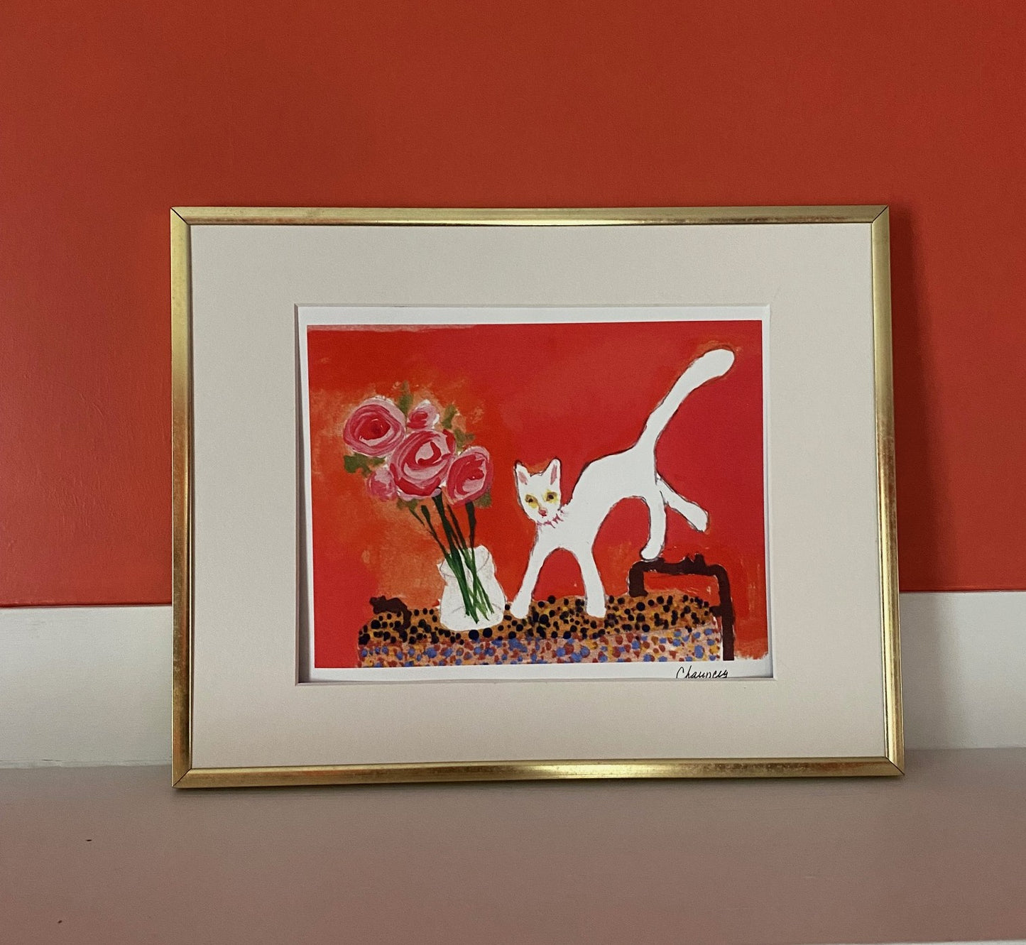 Caspar with Roses art print is in a gold frame with white matting. The Orangey-pink background highlights the white of the cat who has jumped on the table to be near the vase of pink roses.
