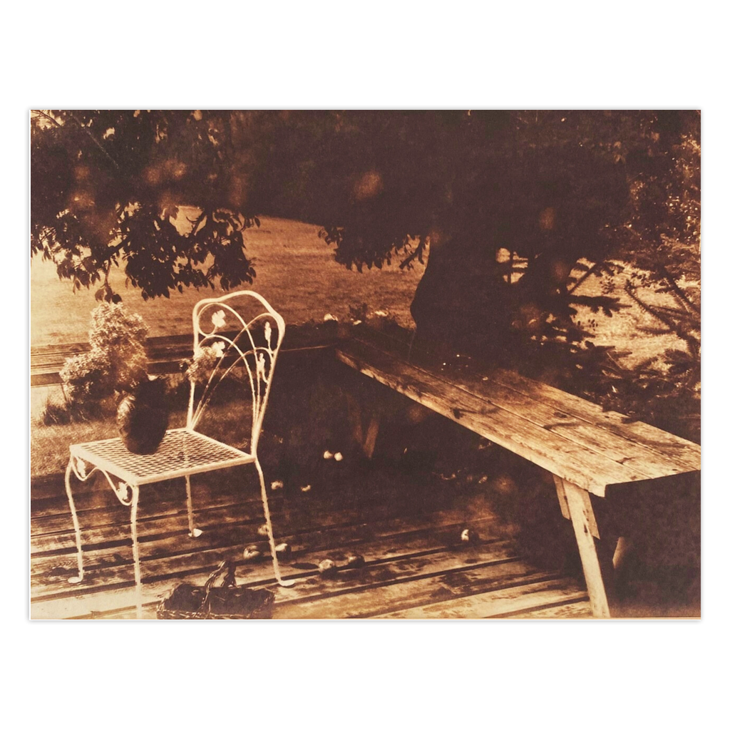 Apples On The Deck is a sepia toned photography card featuring an apple tree that overhangs a wooden deck. A wrought iron chair in white supports a vase with dried flowers.