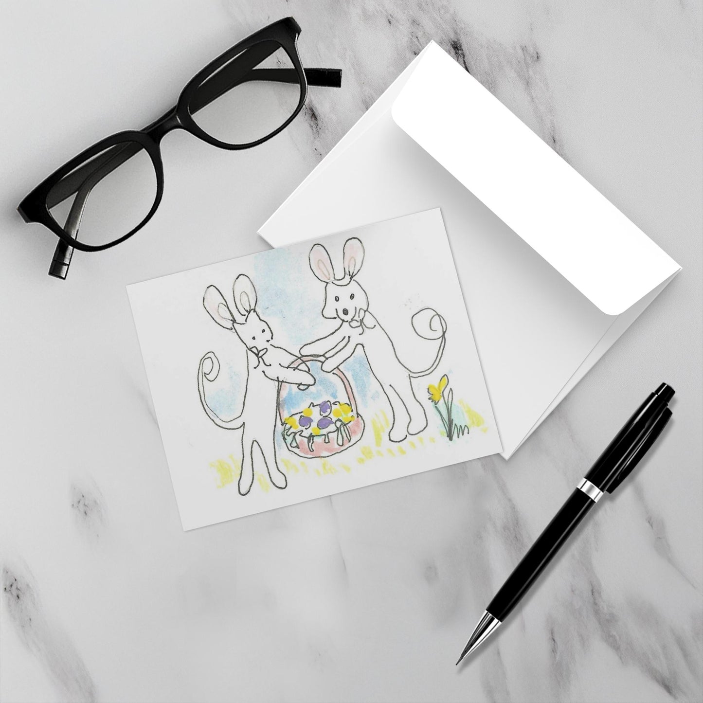 Mockup of Easter Basket From Caspar And Oliver showing card lying on marble surface with black glasses at an angle in the upper right and black pen at an angle in the lower left. A white envelope lies under the card, slightly open.
