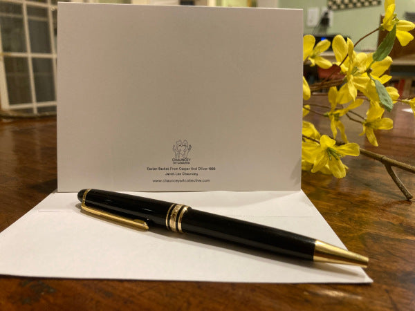 Mockup of the back of the greeting card titled, Easter Basket From Caspar And Oliver standing upright on brown desk with forsythia flowers behind and envelope and black pen in front.