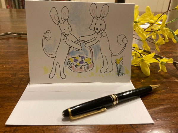 Mockup of the front of the greeting card titled, Easter Basket From Caspar And Oliver standing upright on brown table top with forsythia in the background and white envelope and black pen in the foreground.