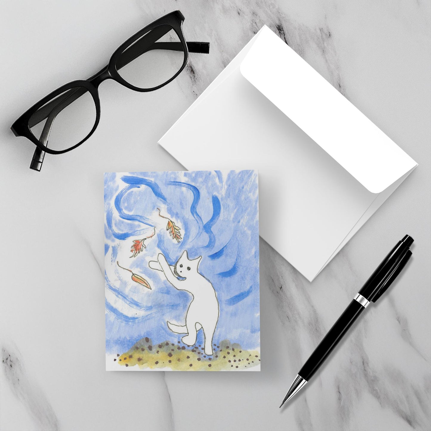 Mockup of the greeting card called Falling Leaves And Caspar which shows the front of the card with the white cat batting three leaves swirling over his head, along side a white envelope, a pair of black reading glasses and a black pen on a grey marble surface.
