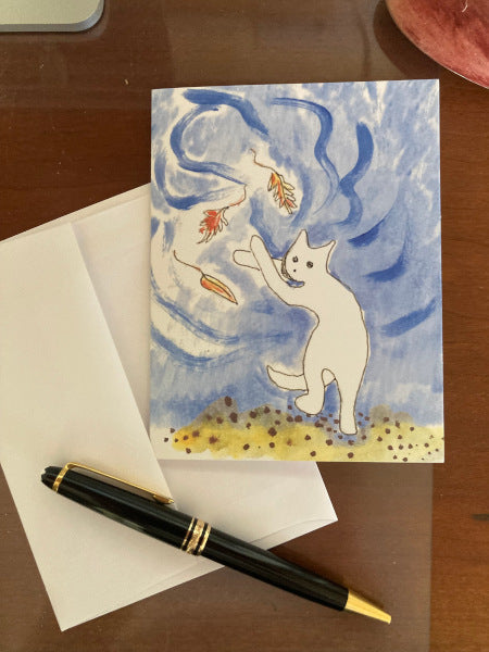 Photograph of the front of the greeting card titled Falling Leaves and Caspar. The card lies atop its white envelope on a brown desk with a black pen at the bottom.