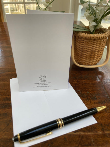 Mockup shows back of notecard and envelope on a wooden table with a fountain pen and a basket filled with white flowers.