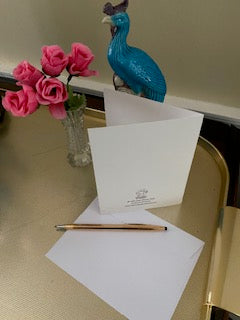 A mock up of the rear of the card on gold tray with miniature pink roses in a vase a blue peacock statuette, the envelope and a gold pen