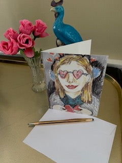 Mock up of the card on a gold tray with pink miniature roses in a vase and a blue peacock statuette. The card is facing the camera on top of the envelope.