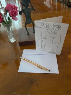 photograph of the front of the greeting card High Top Table For Daddy which is on a brown wooden surface next to an African statuette, a glass vase of pink roses and a white envelope with gold pen.